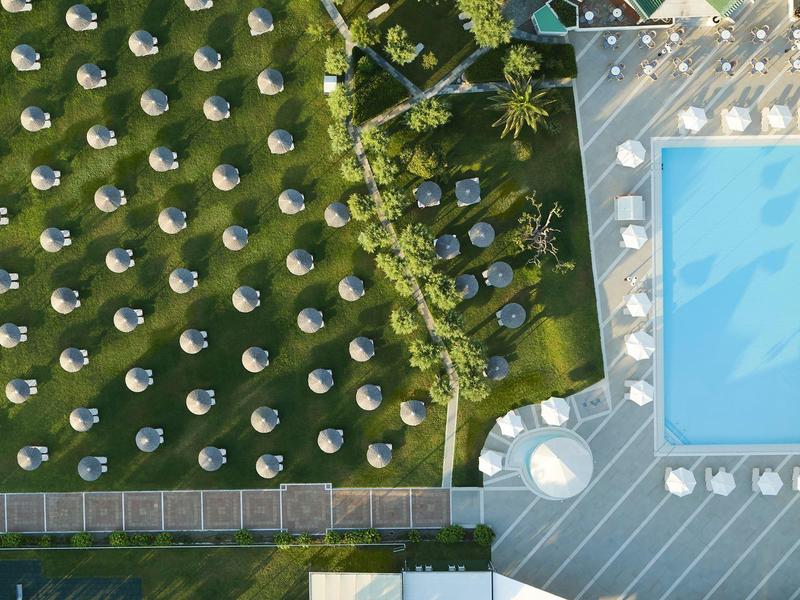 Letecký pohled na bazén vedle trávníku s mnoha bílými slunečníky uspořádanými v řadách.