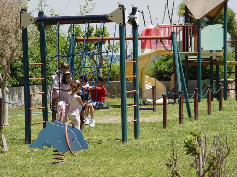 Děti si hrají na prolézačkách a houpačkách v travnatém hřišti.