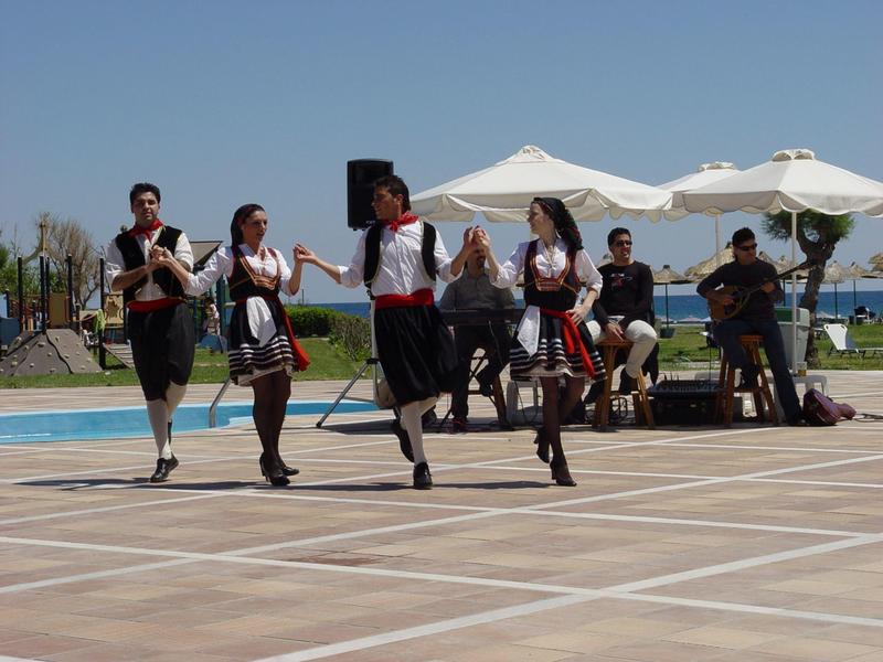 Skupina lidí v tradičních krojích tančí venku na dlážděném prostranství za slunečného počasí.