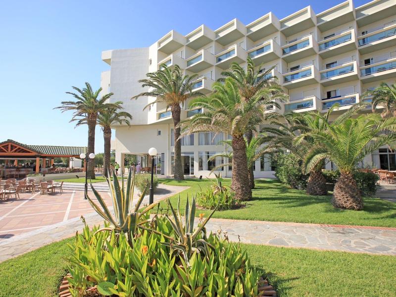 Hotelová budova s balkony, obklopená palmami a zahradou pod jasnou oblohou.