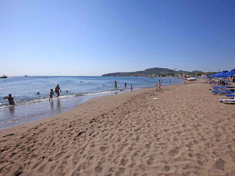 Slunná písečná pláž s lidmi u vody a lehátky pod modrými slunečníky.