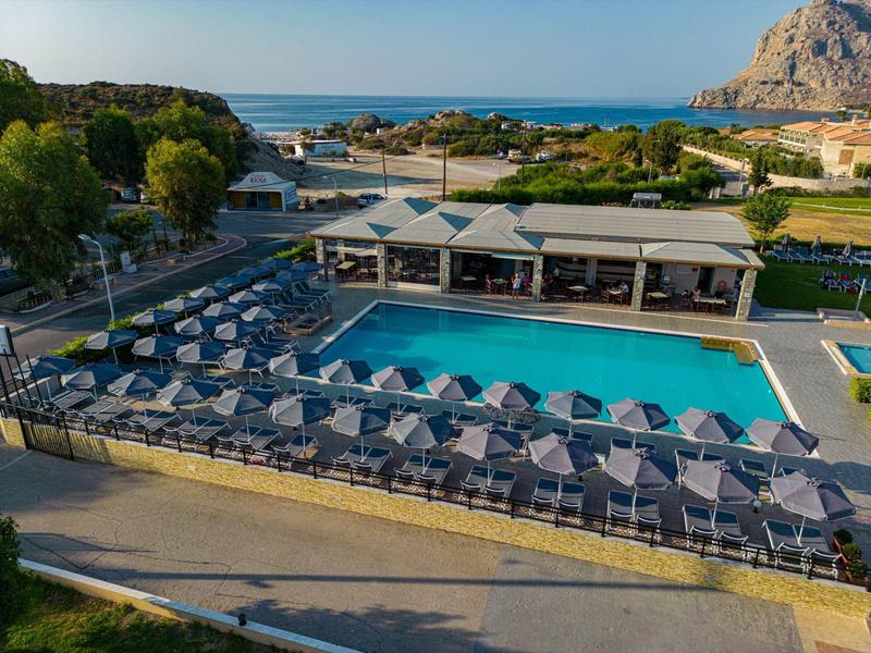 Großer Pool mit vielen blauen Sonnenschirmen, umgeben von Bäumen und Blick aufs Meer im Hintergrund.