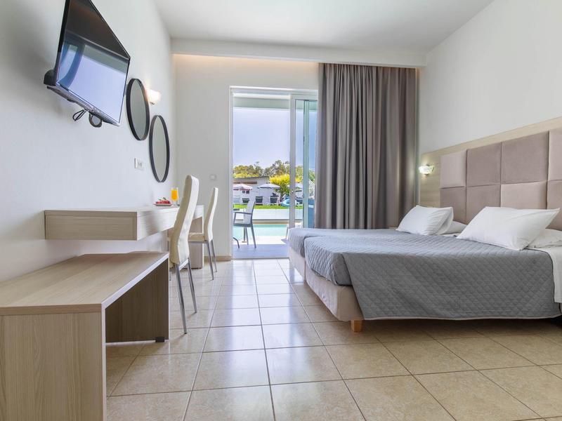 Helles Hotelzimmer mit zwei Betten, Fernseher, Schreibtisch, Stühlen und Balkon mit Blick nach draußen.