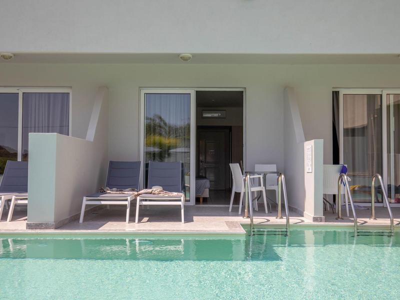 Ein moderner Pool mit klar grünem Wasser vor einer weißen Terrasse mit Liegestühlen und Tischen.