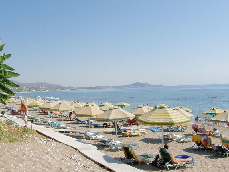 Strand mit Sonnenschirmen, Liegen und wenigen Menschen, blaues Meer, sonniger Himmel, Pflanzen am Rand.