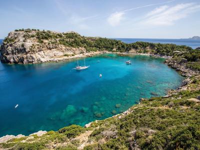 Clear blue water in a sheltered bay with sailboats and green-covered cliffs.