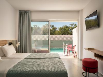 Camera d'hotel luminosa con letto matrimoniale, copriletto verde e balcone con vista piscina.
