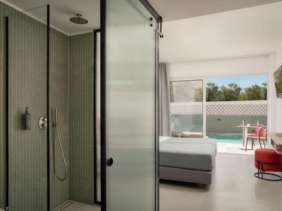 Camera d'hotel moderna con doccia in vetro, letto matrimoniale e vista sulla terrazza con piscina.