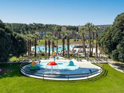 Piscina rotonda con ombrelloni colorati davanti a una grande piscina dell'hotel e palme sotto un cielo azzurro.