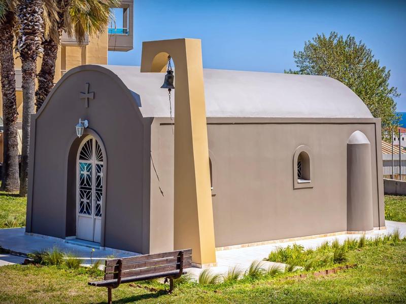 Piccola cappella affascinante con pareti grigio chiaro e porta ad arco, circondata da palme.