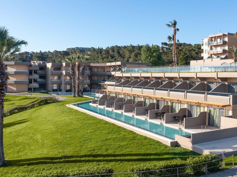 Complesso alberghiero moderno con camere basse, piscine private e prati verdi sotto un cielo azzurro.