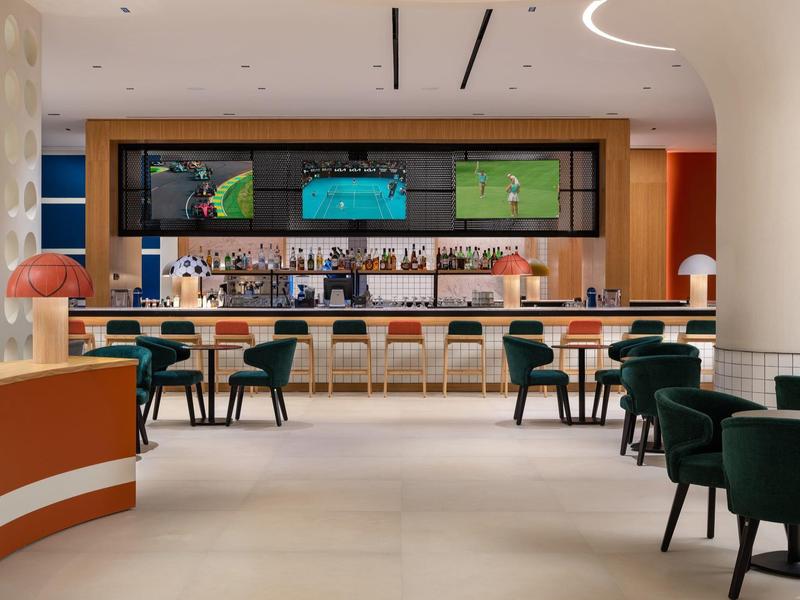 Bar dell'hotel moderna con legno scuro, sedie verdi e trasmissioni sportive sugli schermi.