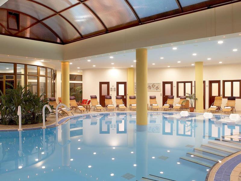 Vnitřní bazén se skleněnou střechou, lehátky a teplým osvětlením v hotelovém prostředí.