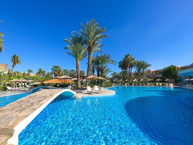 Slunečný rezortní bazén s palmami, čistou modrou vodou a lehátky pod slunečníky.