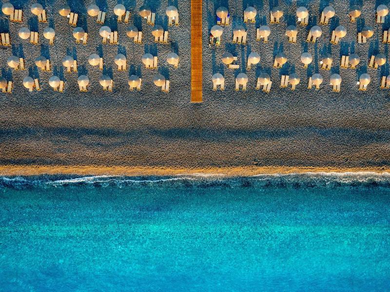 Letecký pohled na písečnou pláž s řadami slunečníků a lehátek vedle čistého modrého moře.