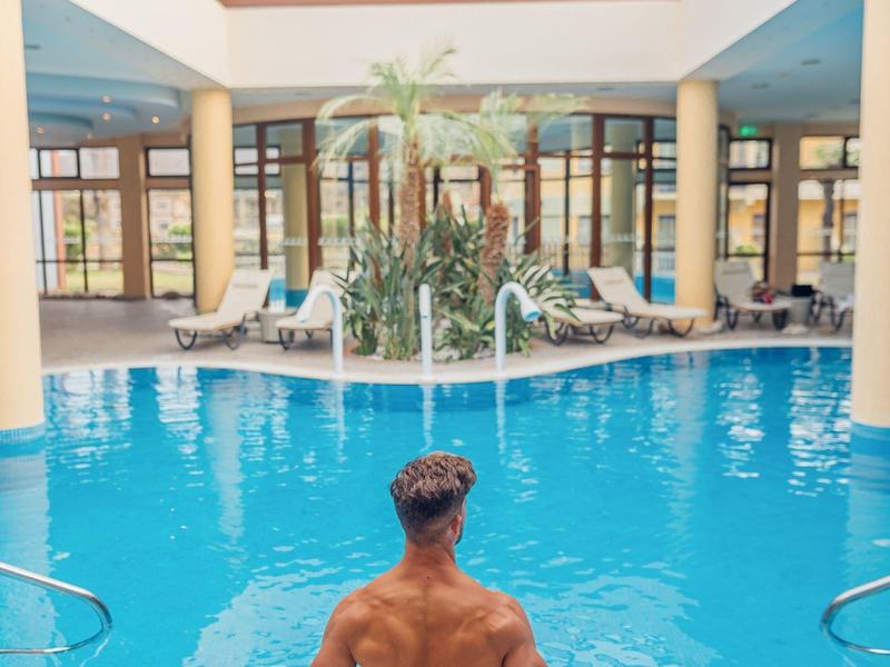 Muž sedí u vnitřního bazénu se skleněnou střechou a lehátky