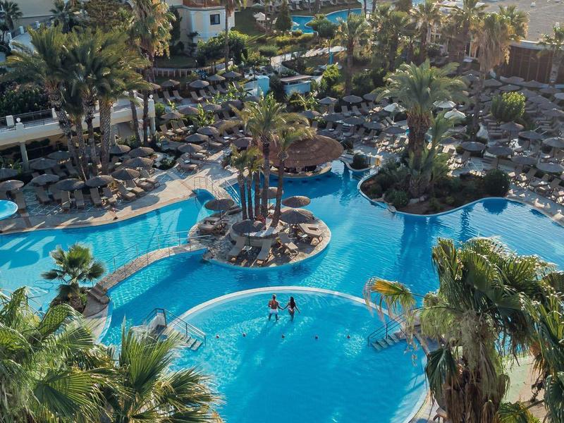 Letecký pohled na resort s lagunovým bazénem, palmami a lehátky u pláže.