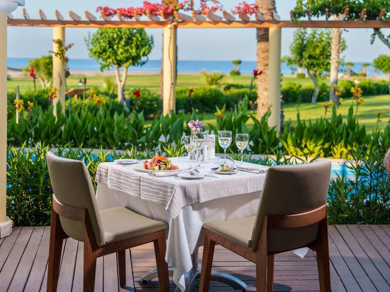 Venkovní jídelní stůl s bílým ubrusem, sklenicemi a květinami u zahrady s výhledem na moře.