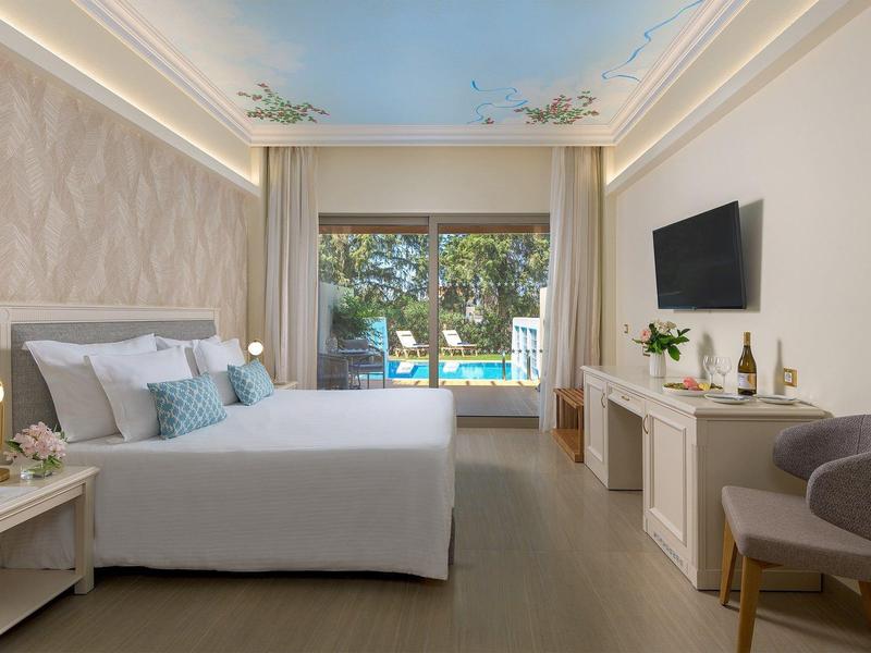 Moderní hotelový pokoj s manželskou postelí, TV, pohodlným křeslem a velkým oknem s výhledem na bazén.