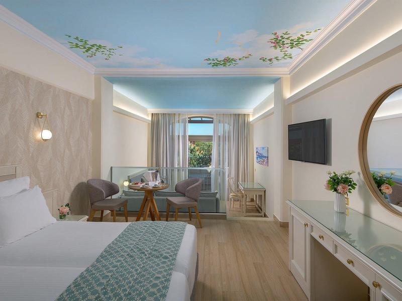 Světý hotelový pokoj s velkou postelí, posezením, dřevěnou podlahou a stropem s přírodním motivem.