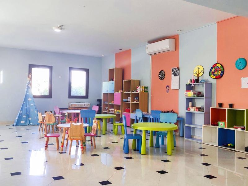 Jasná dětská herna s barevnými stoly, židlemi a hračkami.