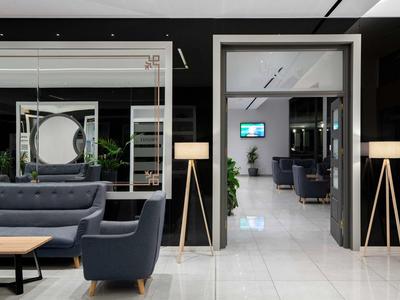 Área de lounge moderna de hotel con sillones grises, sofás y lámparas de pie sobre suelo blanco.