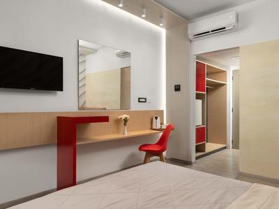 Camera d'hotel moderna con scrivania rossa, sedia, TV a parete e grande specchio con illuminazione LED.