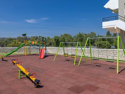 Area giochi esterna con scivoli, altalene e un'altalena a dondolo vicino a un edificio sotto un cielo sereno.