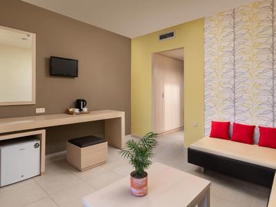 Camera d'hotel moderna con pareti beige, scrivania, TV a schermo piatto, mini frigo, divano con cuscini rossi.