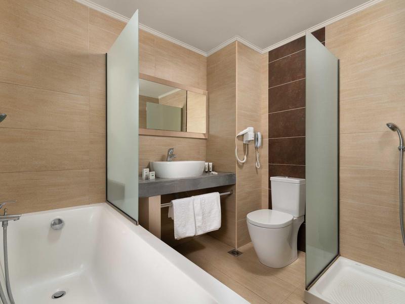 Bagno moderno in hotel con vasca, docce, wc, lavabo e pareti rivestite beige.