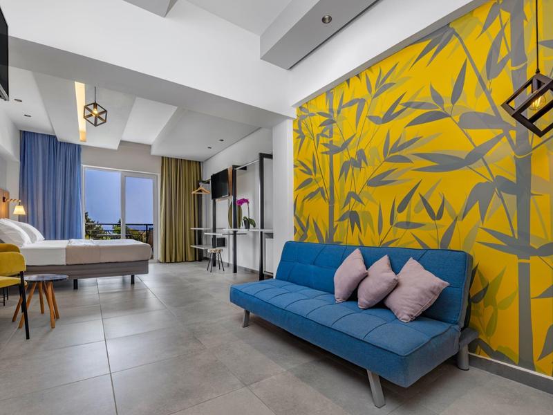 Camera d'hotel luminosa con divano blu, parete gialla decorata, letto e grande finestra con tende.