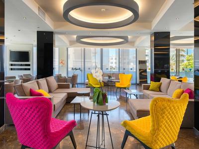 Modern hotel lounge with colorful armchairs in pink, yellow, and brown, and city views through large windows.