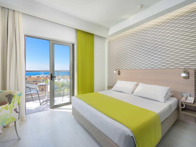 Bright hotel room with double bed, balcony view of the sea, modern decor, and green accents.