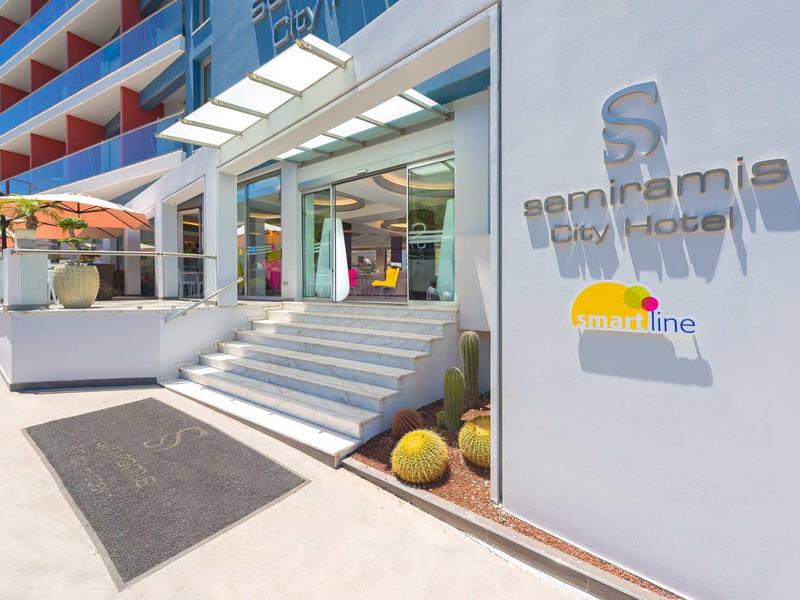 Modern hotel entrance with glass doors, stairs, and decorative plants under a clear sky.