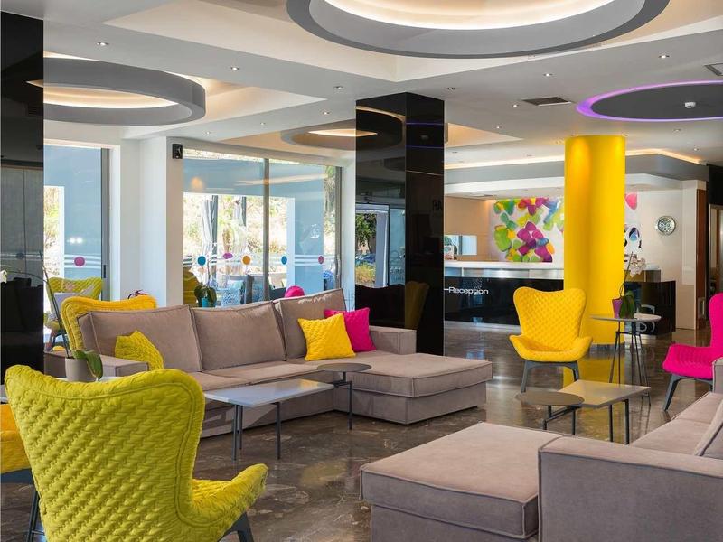 Modern hotel lobby with colorful chairs, gray sofas, circular ceiling lights, and stone floor.