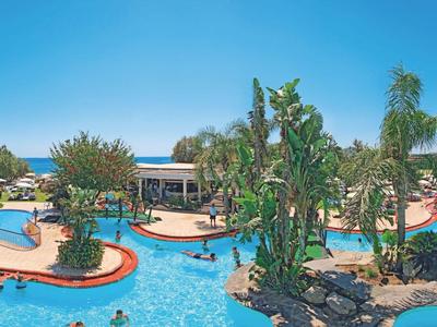 Vue large d'une piscine de station tropicale avec des palmiers, des chaises longues et un ciel dégagé.
