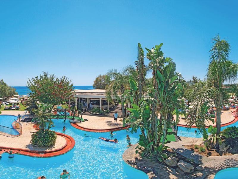 Grande piscina dell'hotel con piante tropicali, palme e lettini sotto un cielo limpido.