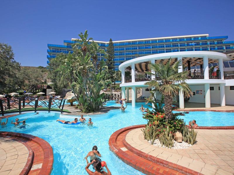 Una piscina d'hotel dal design giocoso, circondata da palme e ombrelloni sotto un cielo azzurro.