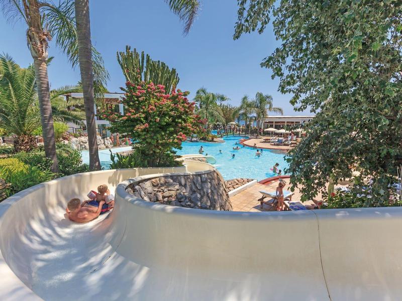Scivolo d'acqua accanto alla piscina con persone sdraiate e vegetazione tropicale in hotel.