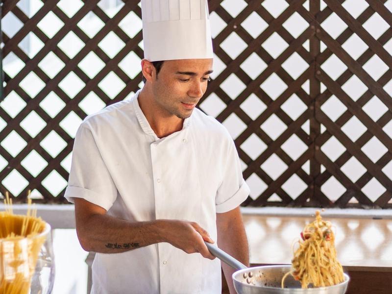 Kucharz w białym uniformie i czapce przygotowuje makaron w kuchni, kolorowe owoce na pierwszym planie.
