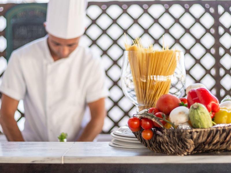 Kucharz w białym ubraniu stoi za stołem z świeżymi warzywami i spaghetti.