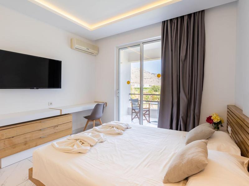 Jasny pokój hotelowy z łóżkiem podwójnym, telewizorem na ścianie i dostępem do balkonu z meblami ogrodowymi.