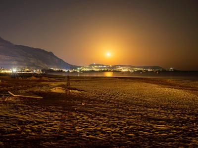 Night beach with illuminated city and mountains in the background under full moon.