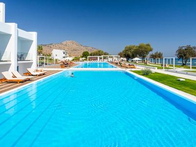 Gran piscina al aire libre con tumbonas, edificio blanco del hotel y cielo despejado.