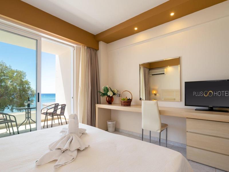 Bright hotel room with double bed, desk, TV, and balcony with sea view.