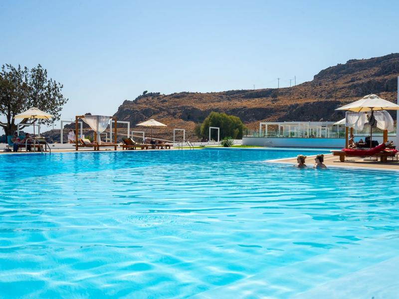 Large hotel pool with sun loungers and hill view under clear blue sky.