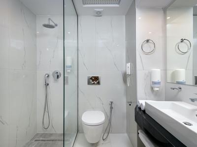 Modern hotel bathroom with glass shower, toilet, and double sink in light colors.