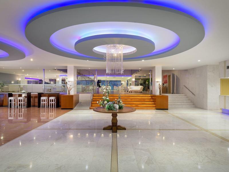 Hall d'hotel ben illuminato con luce circolare sul soffitto e tavolo centrale con composizione floreale.