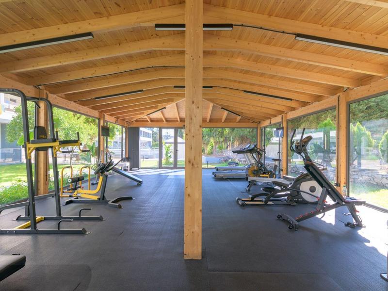 Area fitness coperta con tetto in legno, vari attrezzi da palestra e grandi finestre con vista sul verde