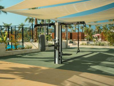 Tende ombreggianti sopra un campo sportivo con attrezzature per esercizi e palme sullo sfondo.